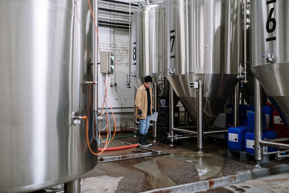 An industrial worker inspects fermentation tanks in a brewery, surrounded by equipment and machinery.