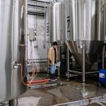 An industrial worker inspects fermentation tanks in a brewery, surrounded by equipment and machinery.