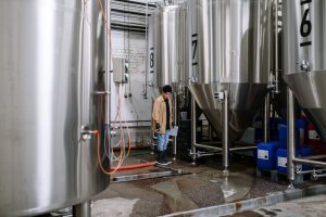 An industrial worker inspects fermentation tanks in a brewery, surrounded by equipment and machinery.